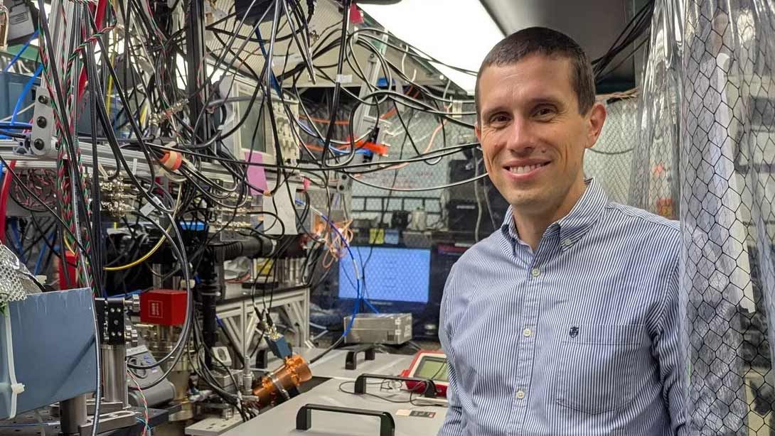 A man in a collared shirt stands in front of lab equipment and many hanging cables.
