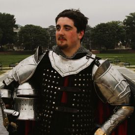 A young man in medieval armour stands proudly, holding his helmet.