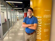 Michael Gullans posing in a modern glass hallway next to a yellow pillar printed with tech terms like "autonomous robotics," "deep learning," and "DISCOVERY."